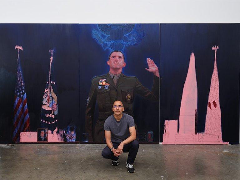 Vincent Valdez crouches in front of a large painting. Valdez is bald and wears glasses, a gray T-shirt and dark pants.